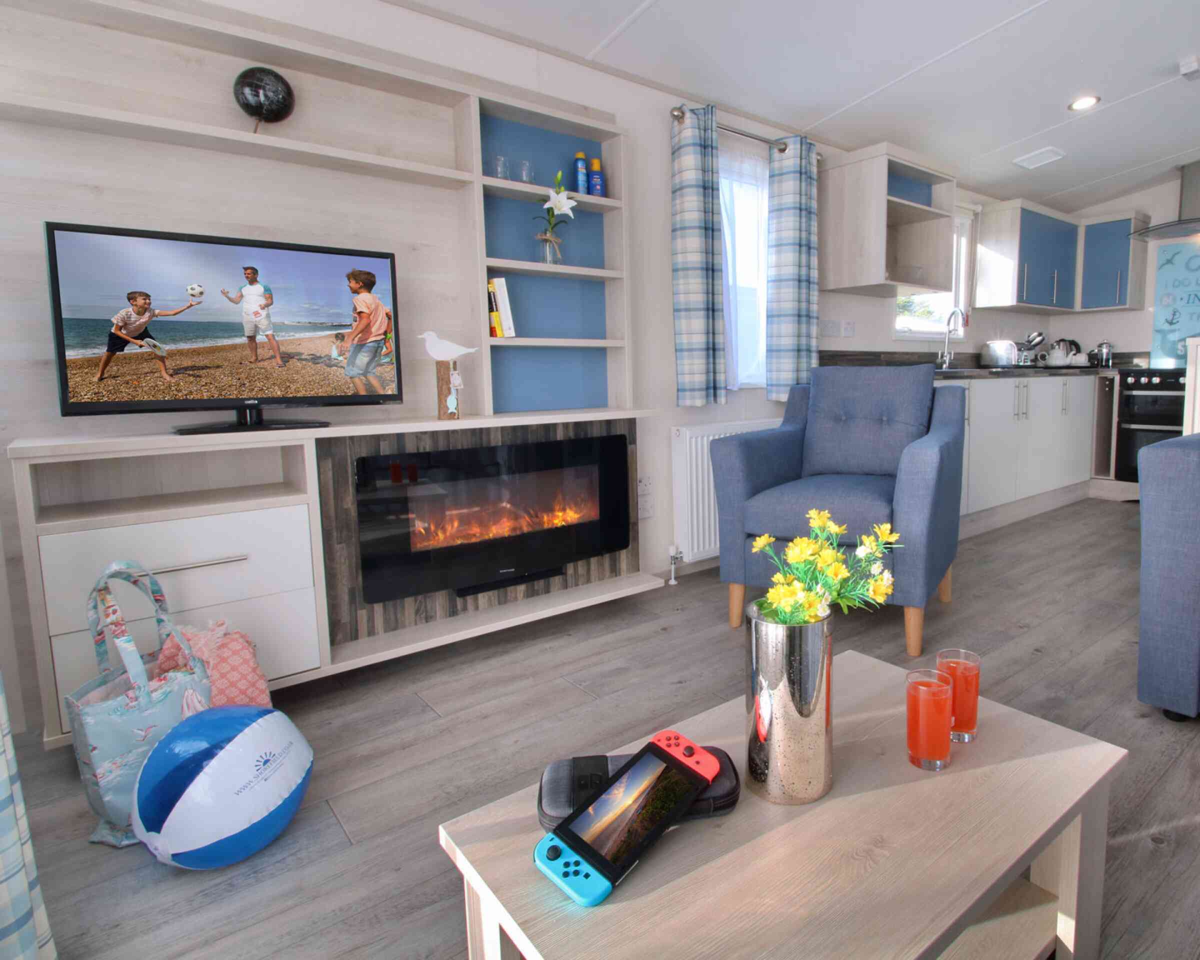 A cozy living area with a stylish light wood entertainment unit featuring a TV displaying children playing beach volleyball. A modern electric fireplace is below the TV. A blue armchair sits beside a table holding a Nintendo Switch and two drinks. A beach ball and a floral arrangement are also present. Bright curtains and a spacious kitchen are visible in the background.