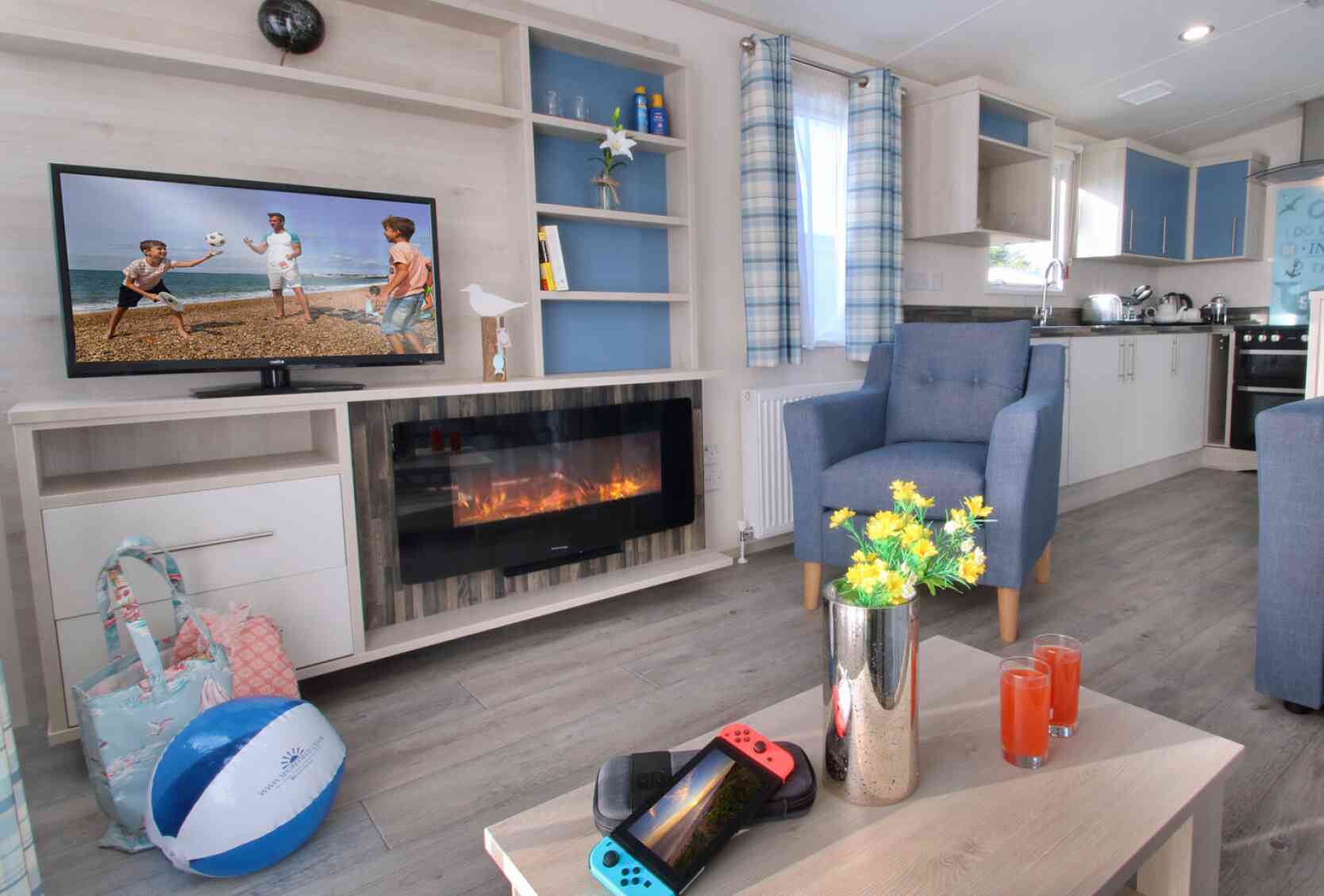 A cozy living area with a stylish light wood entertainment unit featuring a TV displaying children playing beach volleyball. A modern electric fireplace is below the TV. A blue armchair sits beside a table holding a Nintendo Switch and two drinks. A beach ball and a floral arrangement are also present. Bright curtains and a spacious kitchen are visible in the background.