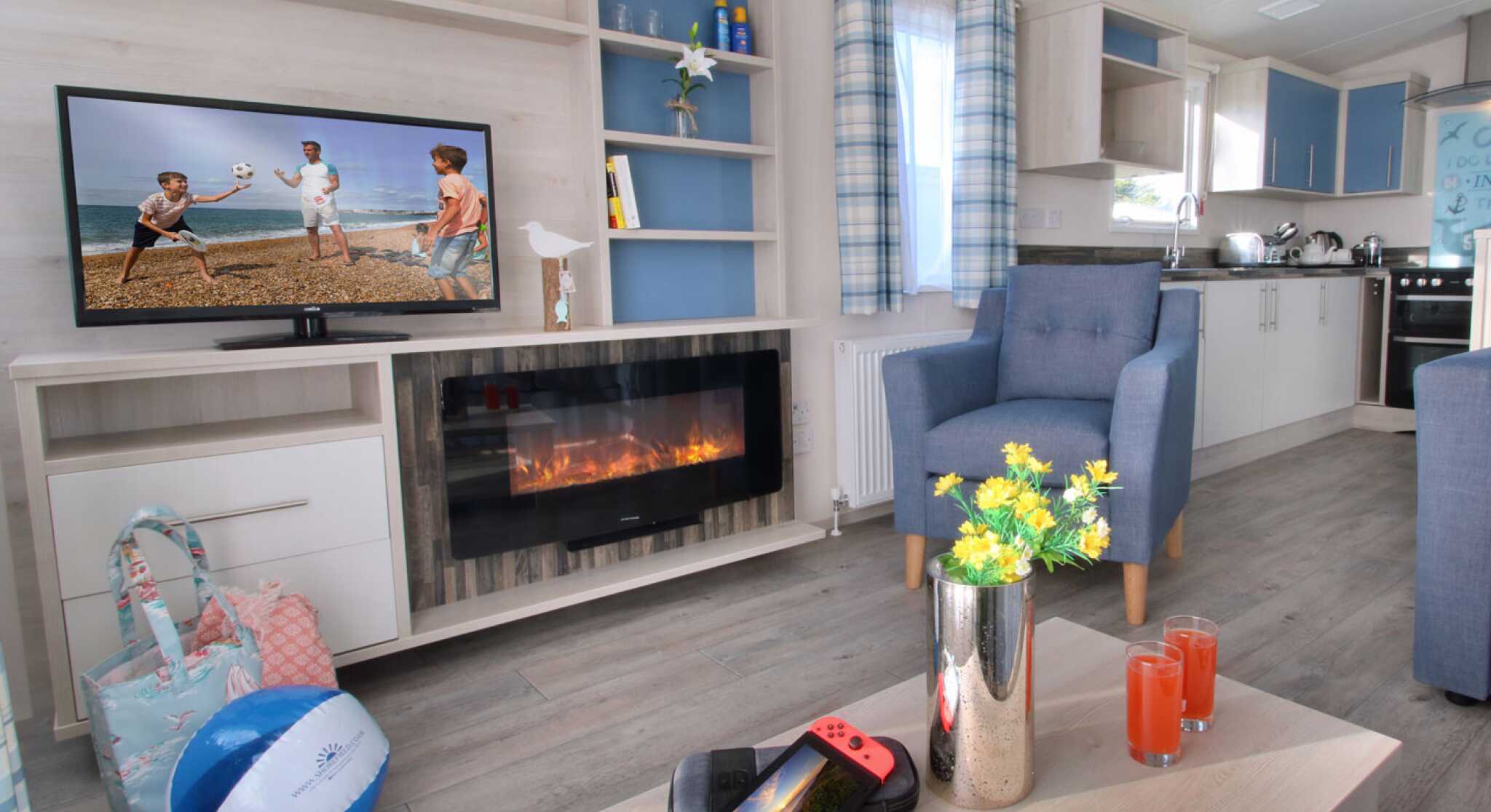 A cozy living area with a stylish light wood entertainment unit featuring a TV displaying children playing beach volleyball. A modern electric fireplace is below the TV. A blue armchair sits beside a table holding a Nintendo Switch and two drinks. A beach ball and a floral arrangement are also present. Bright curtains and a spacious kitchen are visible in the background.