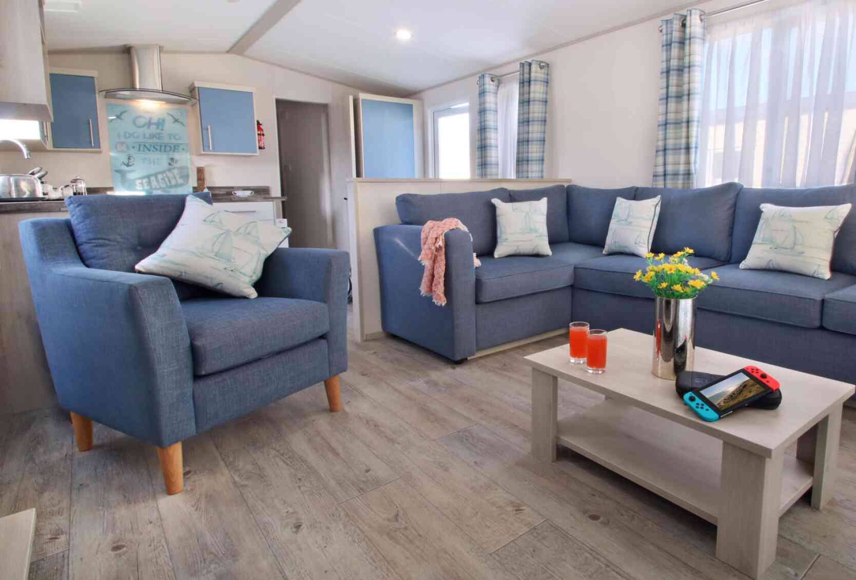 A cozy living room featuring blue armchairs and a sectional sofa adorned with cushions. A light wooden coffee table holds a vase with flowers, two glasses of orange drink, and a Nintendo Switch. Bright curtains frame the windows, enhancing the inviting atmosphere.