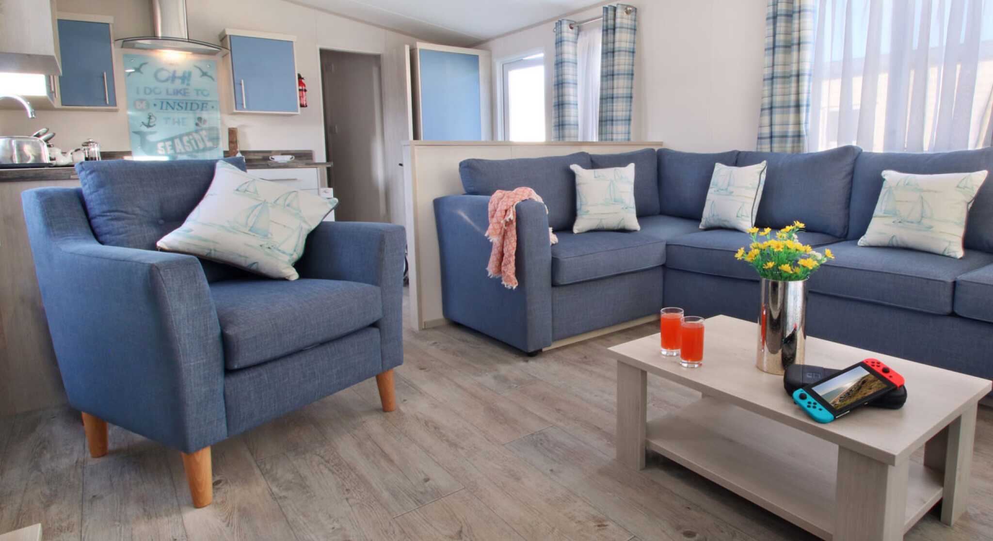A cozy living room featuring blue armchairs and a sectional sofa adorned with cushions. A light wooden coffee table holds a vase with flowers, two glasses of orange drink, and a Nintendo Switch. Bright curtains frame the windows, enhancing the inviting atmosphere.
