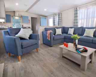 A cozy living room featuring blue armchairs and a sectional sofa adorned with cushions. A light wooden coffee table holds a vase with flowers, two glasses of orange drink, and a Nintendo Switch. Bright curtains frame the windows, enhancing the inviting atmosphere.