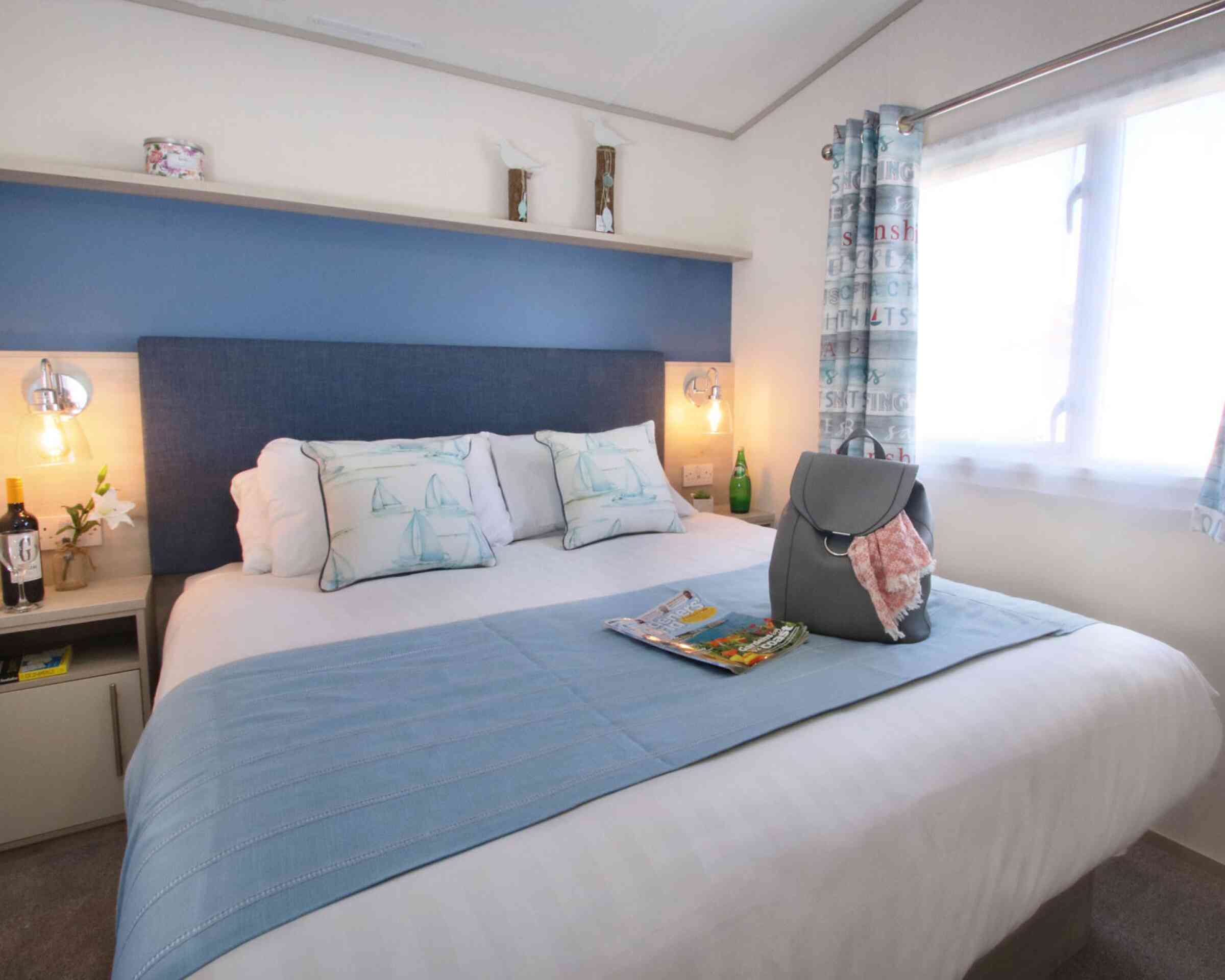 A cozy bedroom featuring a comfortable bed with blue and white accents, decorative pillows, and a light blue throw blanket. A bedside table holds a small bottle and glass, while a handbag and magazine sit on the bed. Natural light filters through a window with patterned curtains.