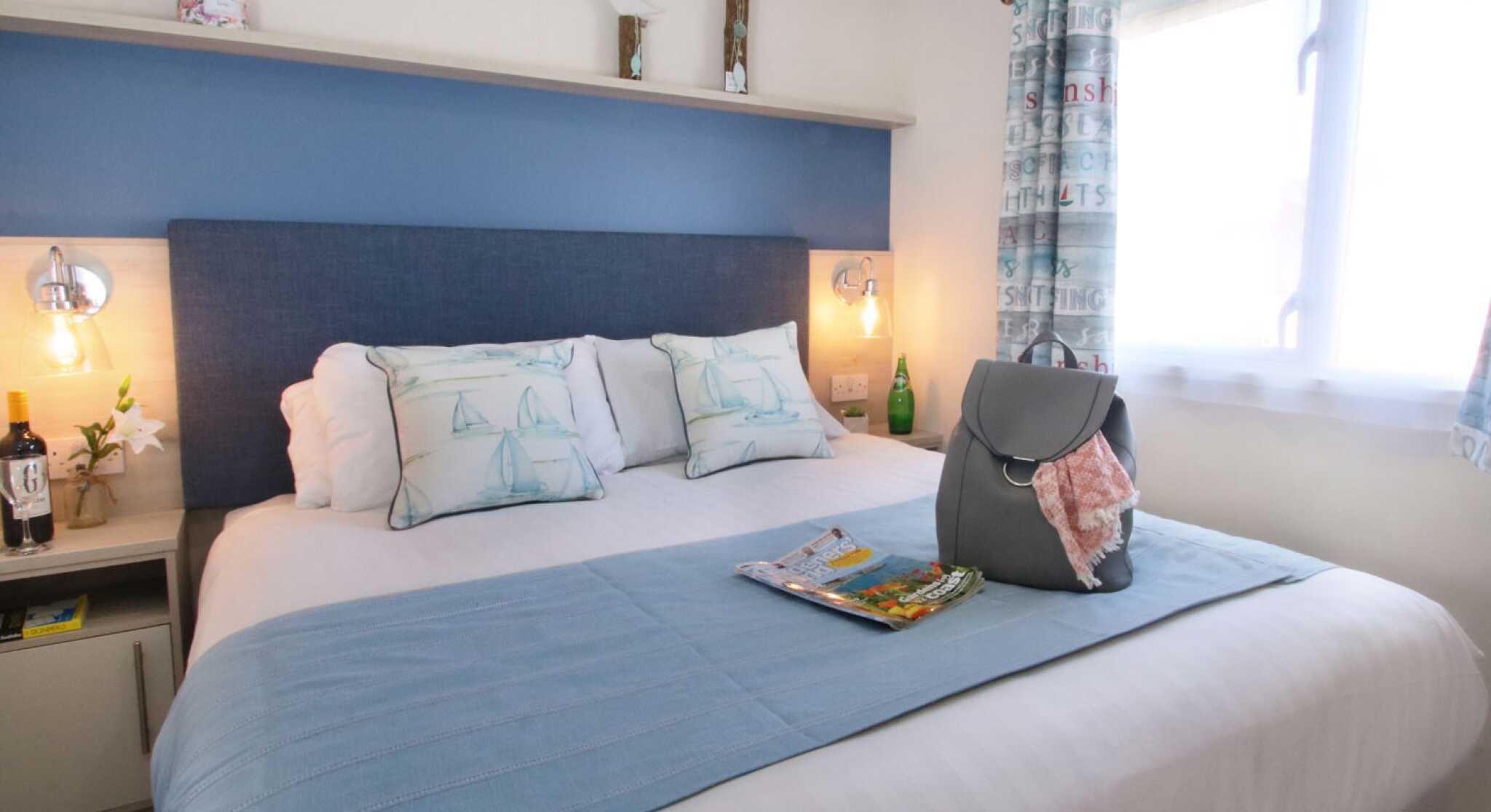 A cozy bedroom featuring a comfortable bed with blue and white accents, decorative pillows, and a light blue throw blanket. A bedside table holds a small bottle and glass, while a handbag and magazine sit on the bed. Natural light filters through a window with patterned curtains.