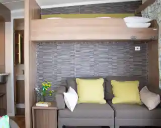 A cozy room featuring a sofa with yellow cushions, a wooden partition with a bunk bed above, and a decorative wall with a textured pattern. A small bouquet of flowers and a bedside table add charm to the space.