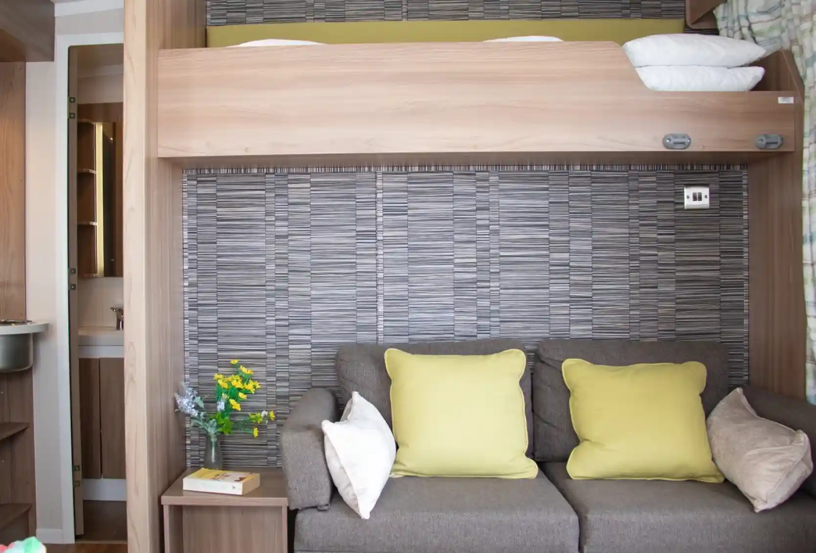 A cozy room featuring a sofa with yellow cushions, a wooden partition with a bunk bed above, and a decorative wall with a textured pattern. A small bouquet of flowers and a bedside table add charm to the space.