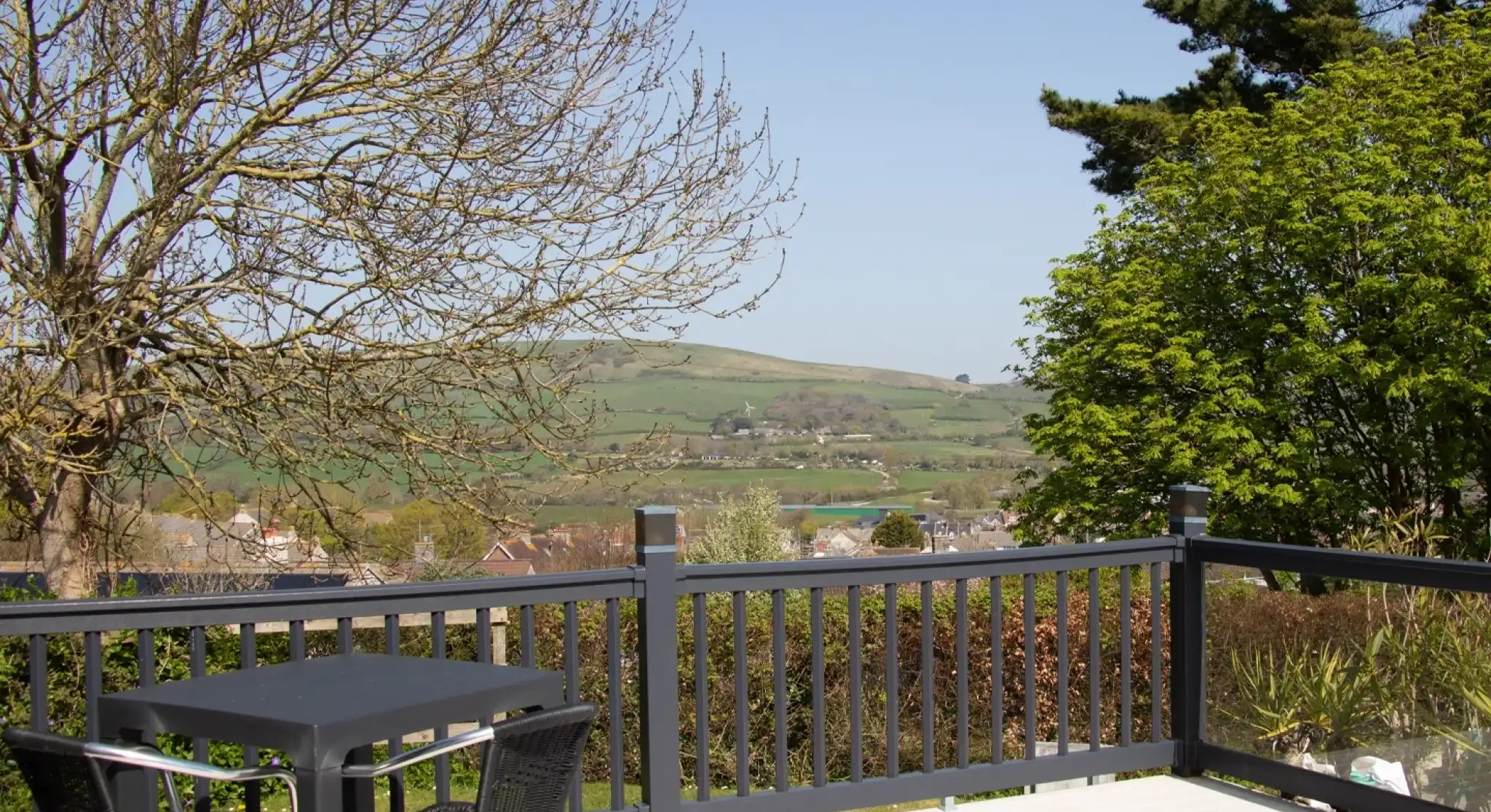 A sunny view from a deck featuring a black table and chairs. In the background, rolling hills and a clear blue sky can be seen, with sparse trees nearby.