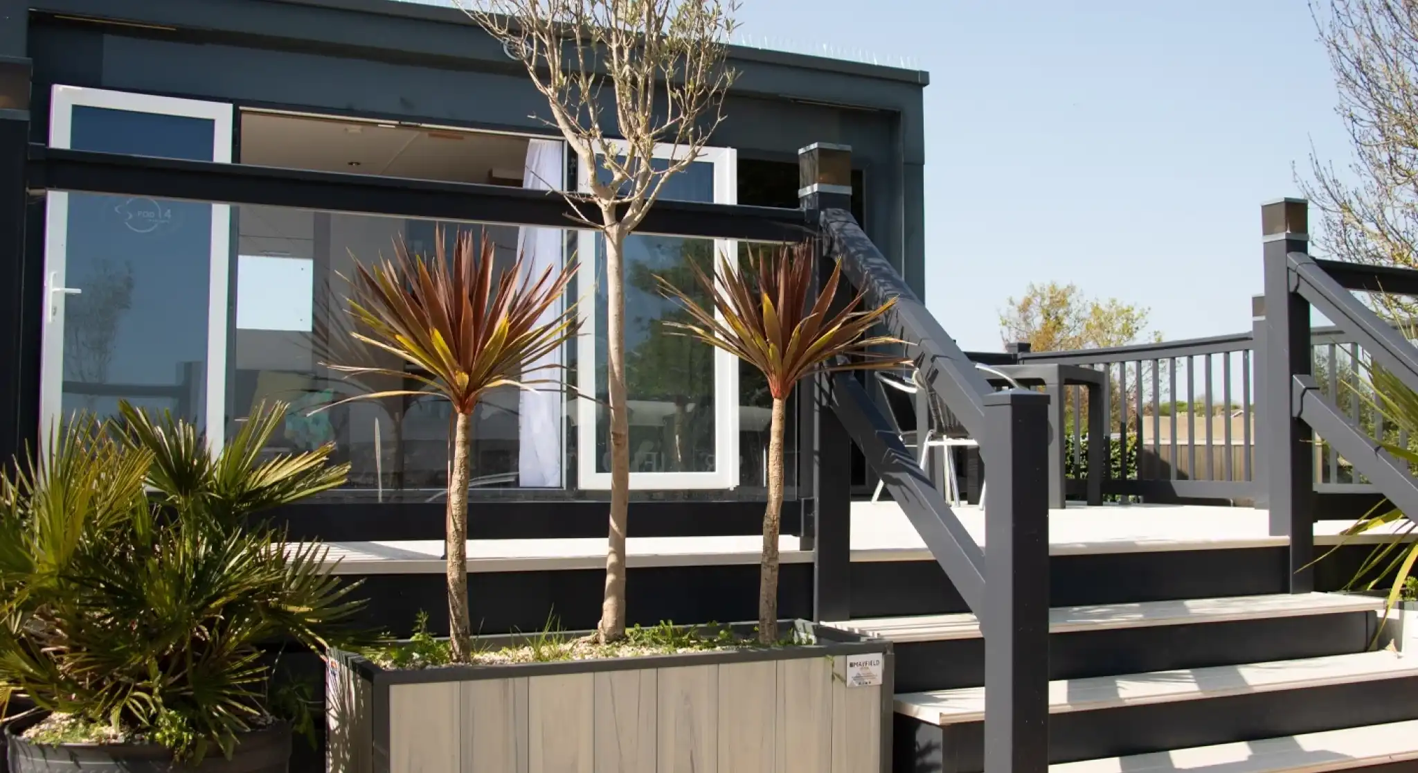 A modern, black and gray outdoor deck featuring a staircase and two potted plants. The background shows a building with large windows, surrounded by greenery and a clear blue sky.