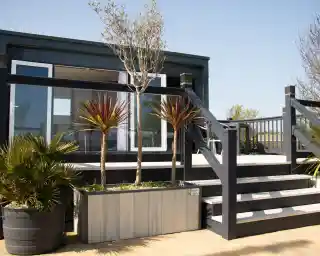 A modern, black and gray outdoor deck featuring a staircase and two potted plants. The background shows a building with large windows, surrounded by greenery and a clear blue sky.