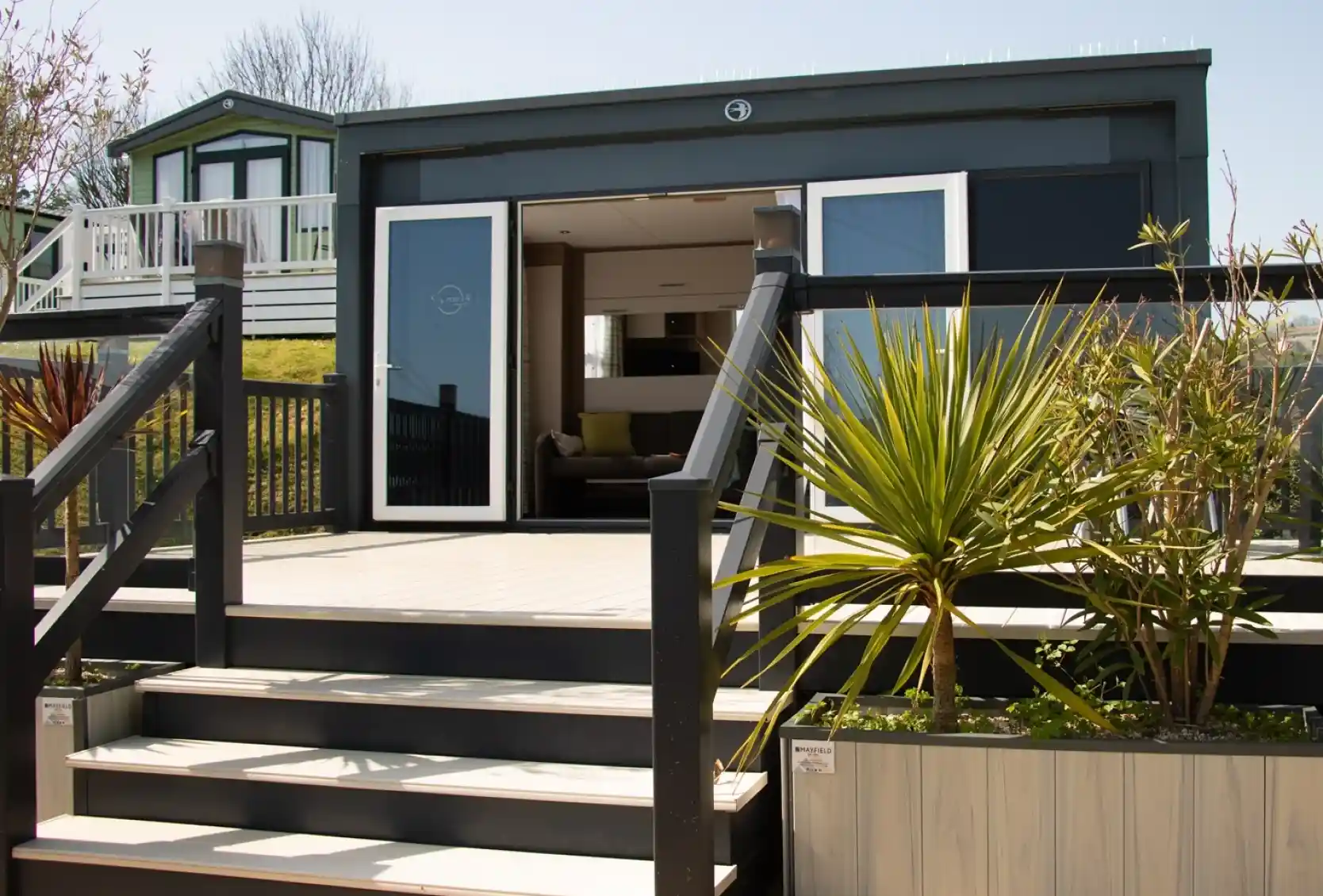 A modern bungalow with a wooden deck and stairs leading up to a large glass door. The surrounding area features plants and a neatly designed garden. In the background, another building can be seen on a slight elevation.