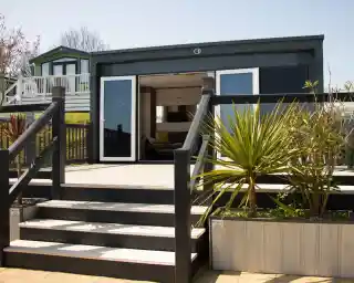 A modern bungalow with a wooden deck and stairs leading up to a large glass door. The surrounding area features plants and a neatly designed garden. In the background, another building can be seen on a slight elevation.