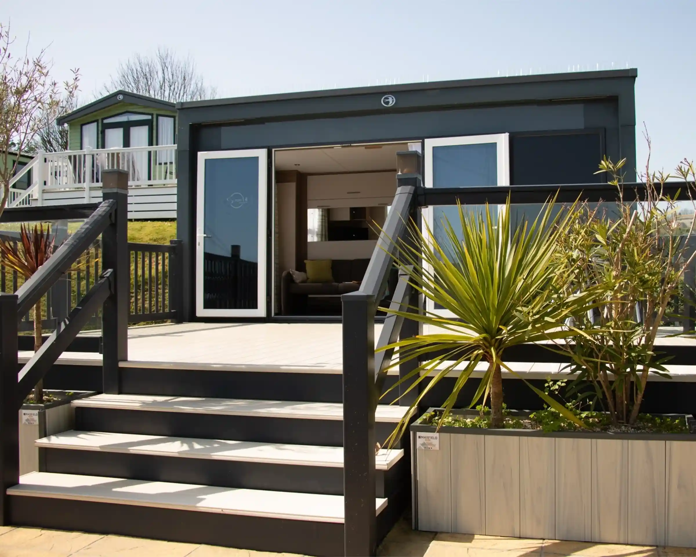 A modern bungalow with a wooden deck and stairs leading up to a large glass door. The surrounding area features plants and a neatly designed garden. In the background, another building can be seen on a slight elevation.
