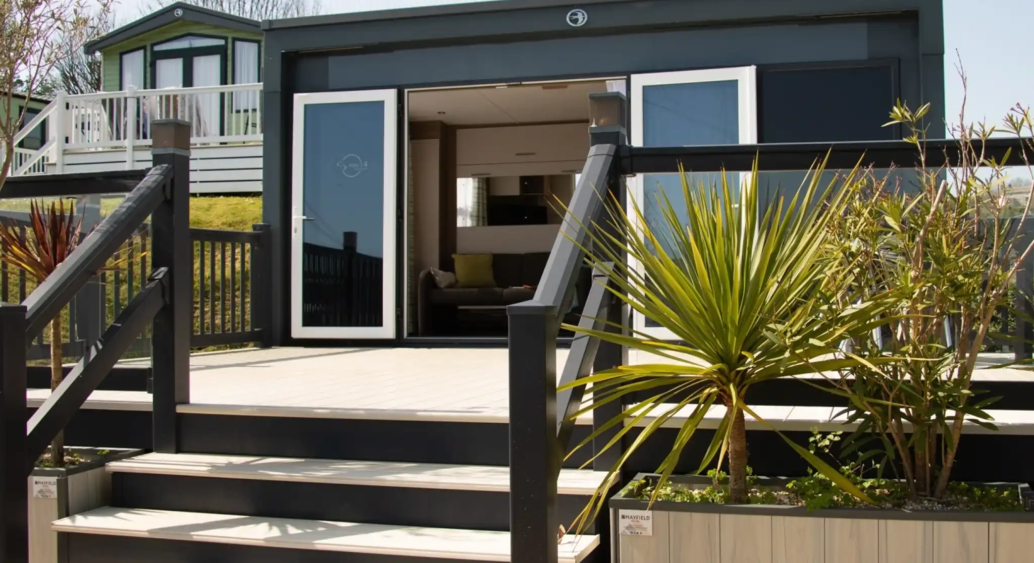 A modern bungalow with a wooden deck and stairs leading up to a large glass door. The surrounding area features plants and a neatly designed garden. In the background, another building can be seen on a slight elevation.