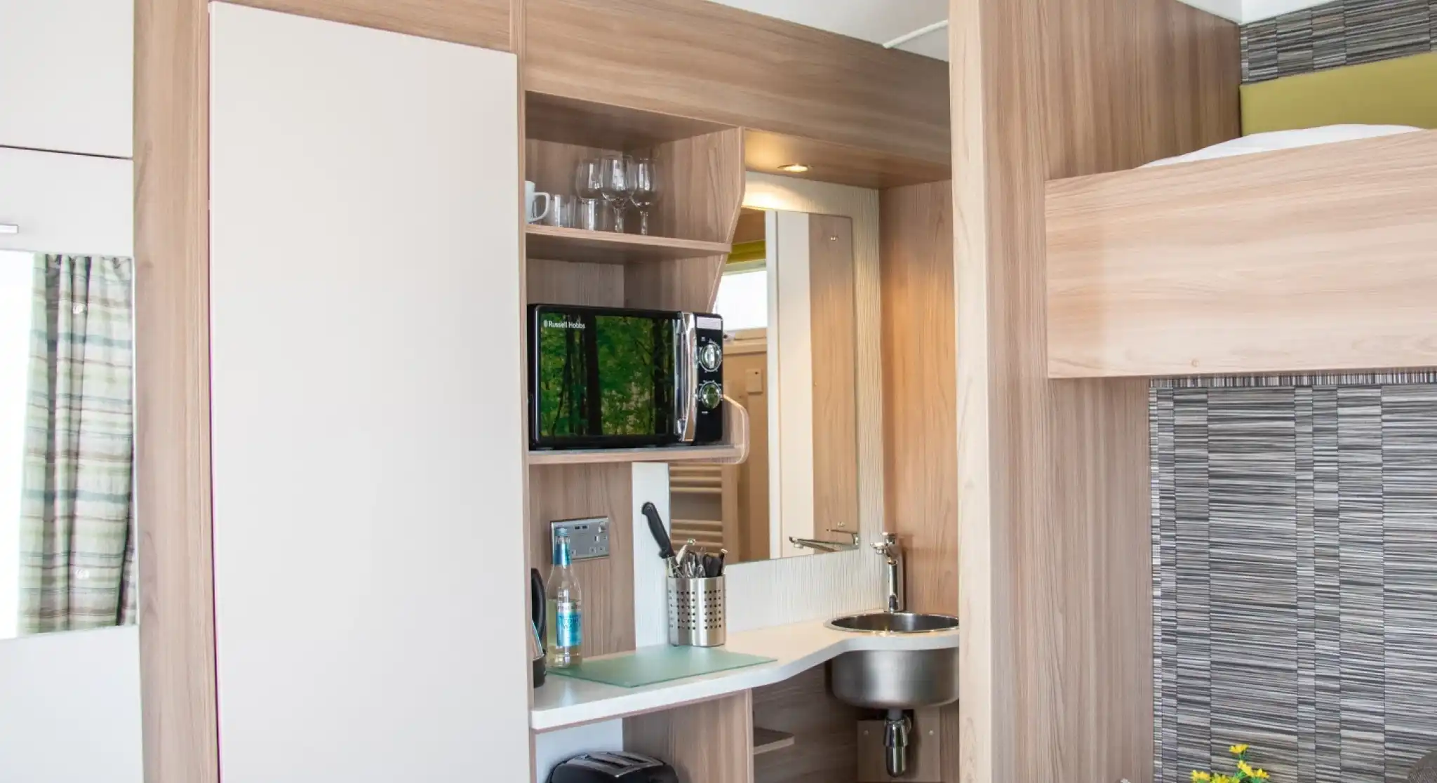 A compact kitchenette with wooden cabinetry features a small sink, microwave, and a shelf with glasses. A cozy sofa is visible beside the kitchen area. Brightly lit space with modern decor.