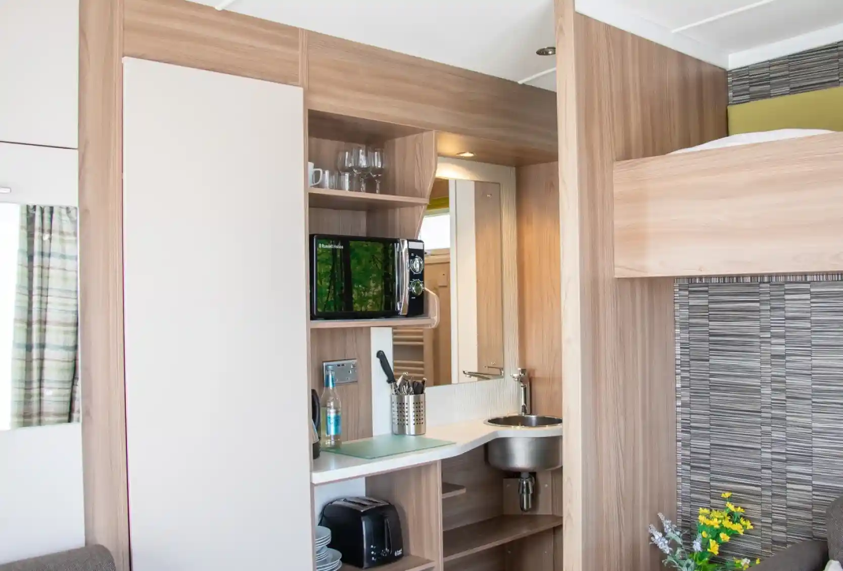 A compact kitchenette with wooden cabinetry features a small sink, microwave, and a shelf with glasses. A cozy sofa is visible beside the kitchen area. Brightly lit space with modern decor.