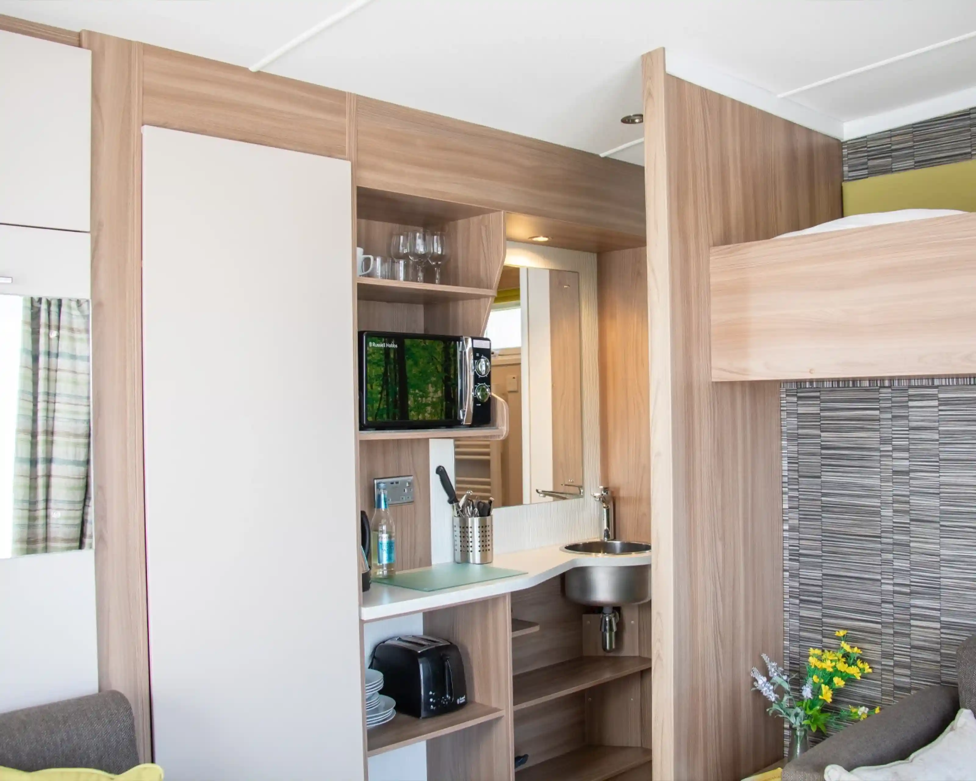 A compact kitchenette with wooden cabinetry features a small sink, microwave, and a shelf with glasses. A cozy sofa is visible beside the kitchen area. Brightly lit space with modern decor.