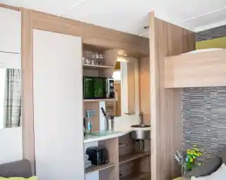 A compact kitchenette with wooden cabinetry features a small sink, microwave, and a shelf with glasses. A cozy sofa is visible beside the kitchen area. Brightly lit space with modern decor.
