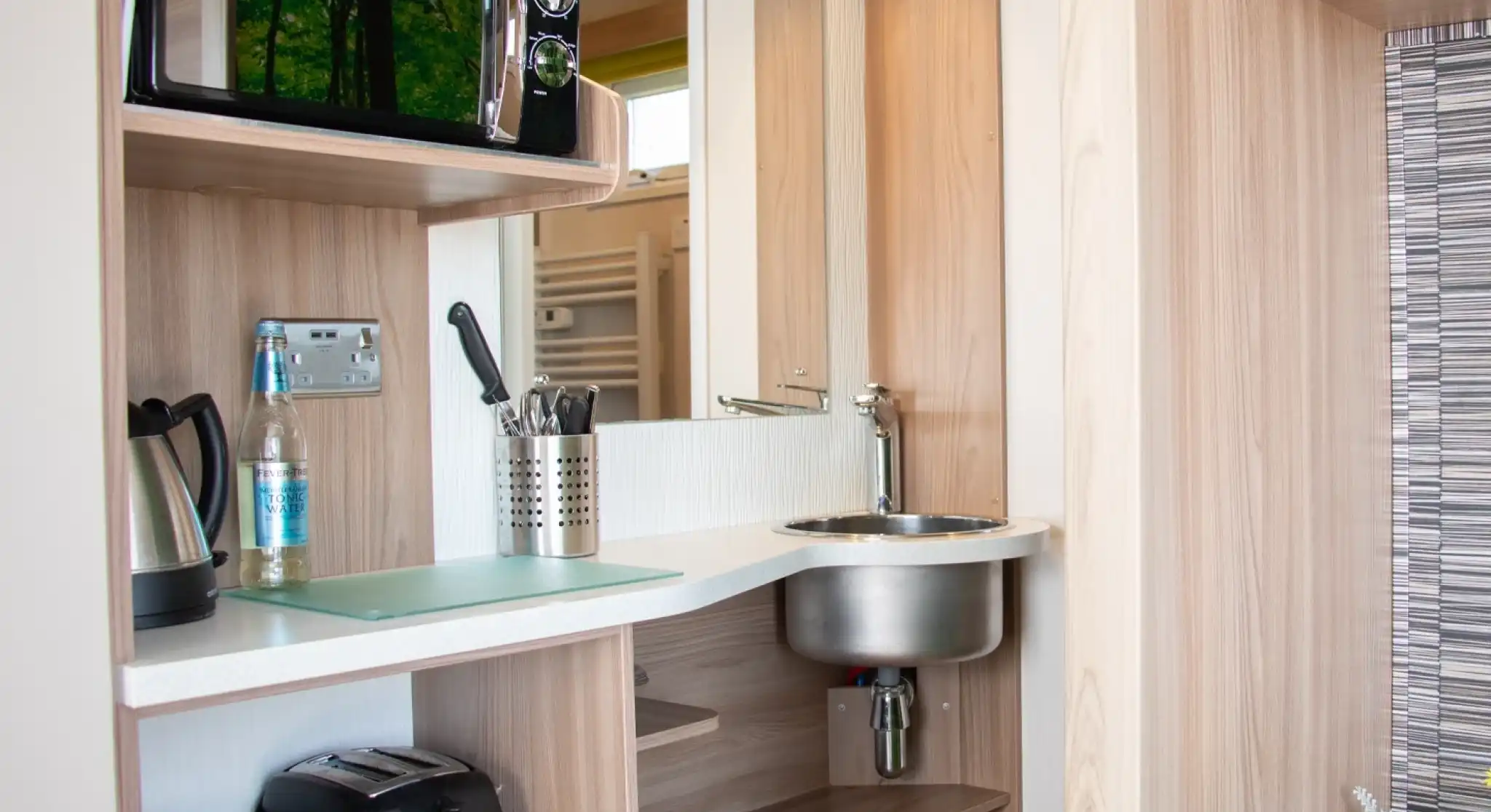 A modern kitchenette featuring a microwave, kettle, sink, and toaster, set against wooden cabinetry. Utensils are neatly arranged near the sink, and a bottle of water sits on the counter.