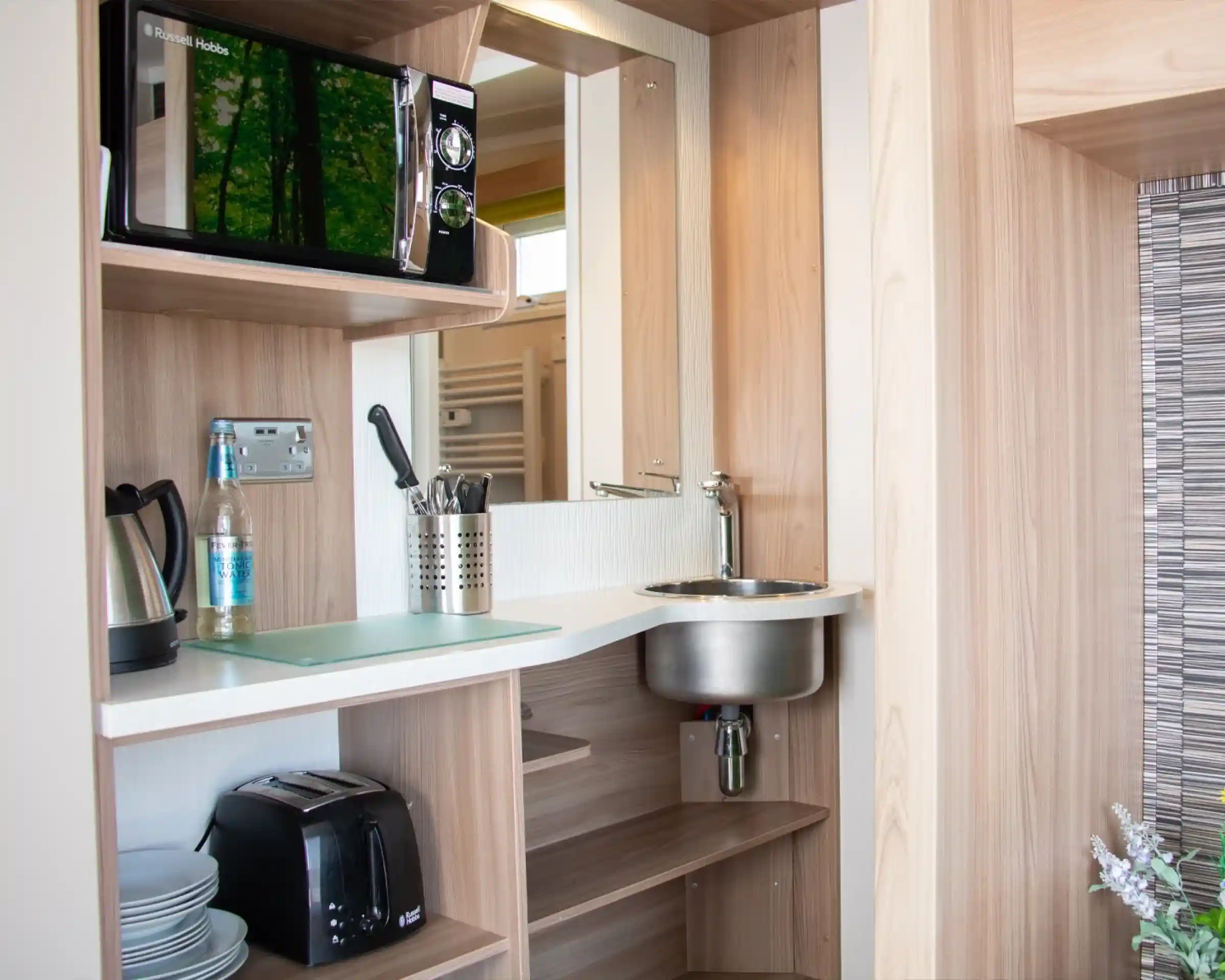 A modern kitchenette featuring a microwave, kettle, sink, and toaster, set against wooden cabinetry. Utensils are neatly arranged near the sink, and a bottle of water sits on the counter.
