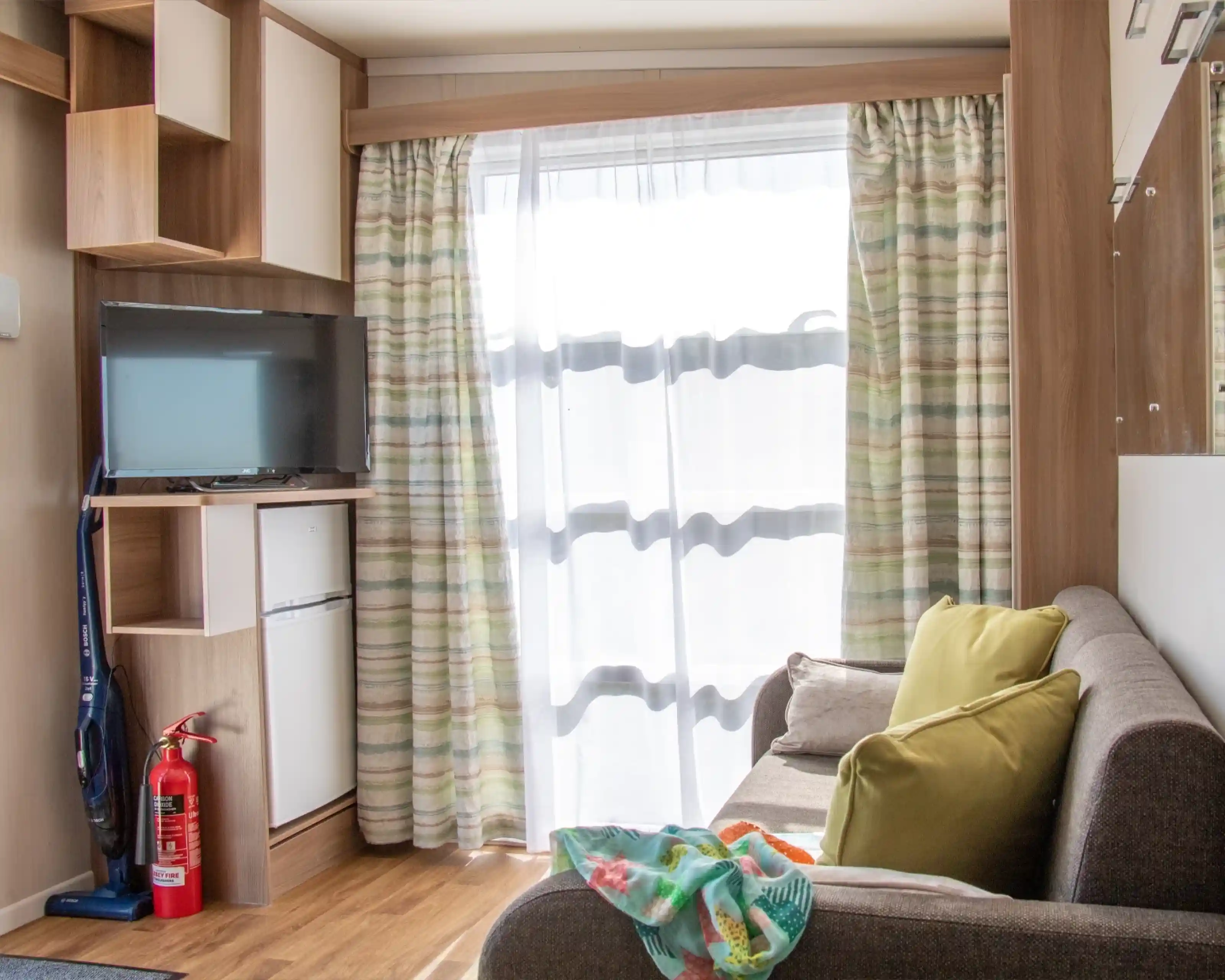 A cozy interior of a small living space featuring a sofa with green and yellow pillows, a television mounted on the wall, and a small white refrigerator. Light streams through sheer curtains, illuminating wooden flooring and built-in shelves. A fire extinguisher is positioned in the corner.