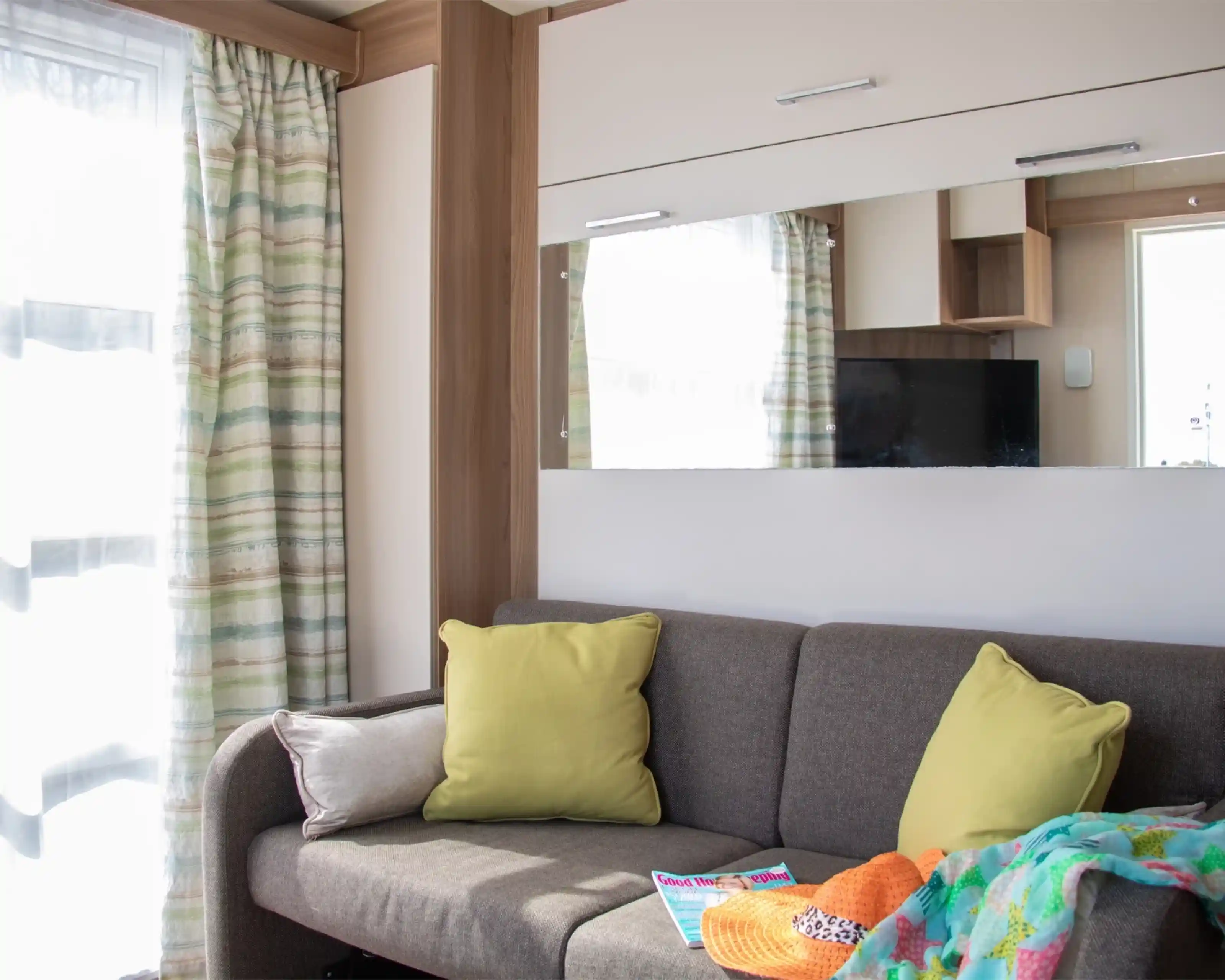A cozy living space featuring a grey sofa adorned with bright yellow cushions. There is a colorful blanket draped over one side, and a magazine resting on the sofa. Light streams in through sheer curtains, with a mirrored wall reflecting the room.