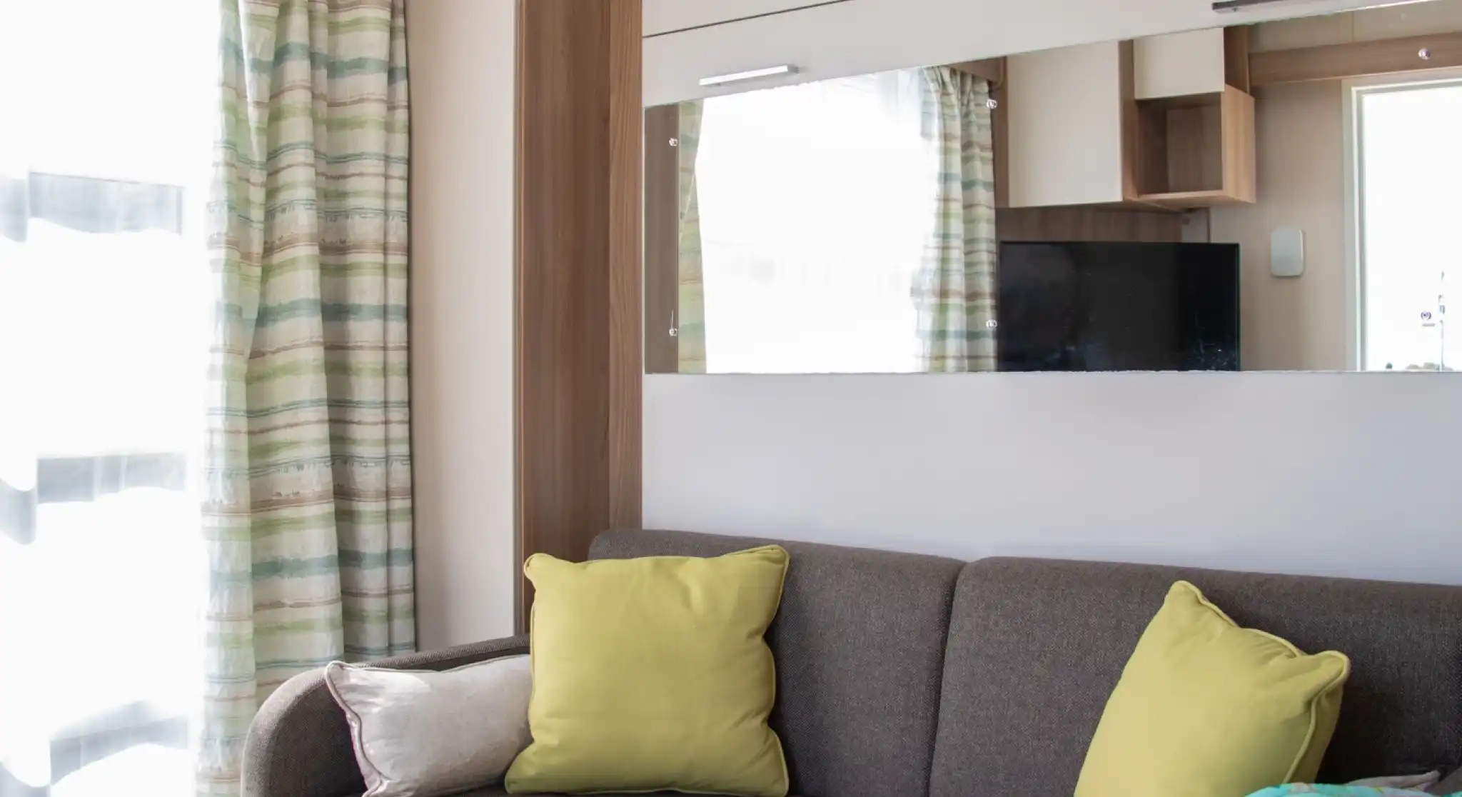A cozy living space featuring a grey sofa adorned with bright yellow cushions. There is a colorful blanket draped over one side, and a magazine resting on the sofa. Light streams in through sheer curtains, with a mirrored wall reflecting the room.