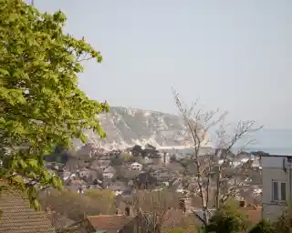 A view of a coastal landscape featuring a green hillside, distant cliffs, and a small town with houses nestled among trees. The sky is clear and blue, enhancing the serene atmosphere.