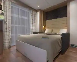 A cozy bedroom featuring a platform bed with a gray and beige color scheme. The backdrop has wood paneling and soft lighting. A small bookshelf is visible with a book resting on the bed. Sheer curtains let in natural light, and the flooring is made of warm-toned wood.