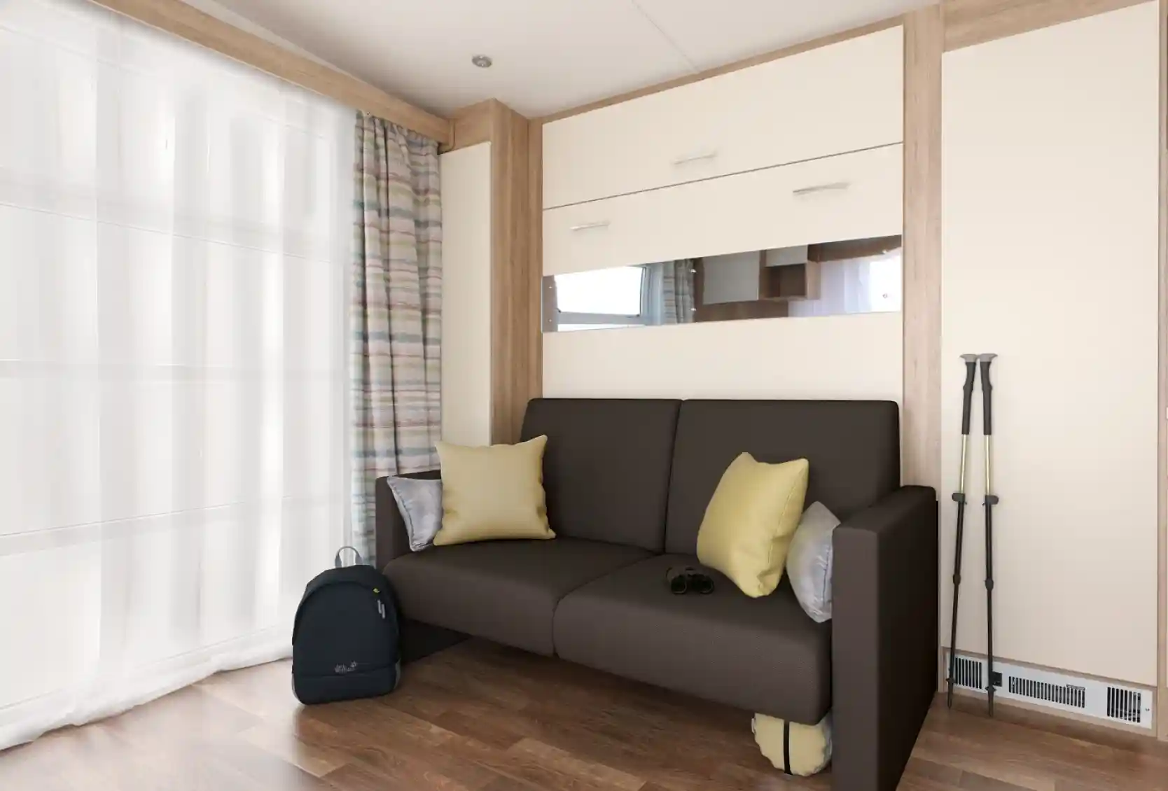 A modern living space featuring a brown sofa with yellow cushions, against a backdrop of light-colored walls and wooden accents. Next to the sofa is a black backpack and a pair of binoculars. Sheer curtains let in natural light, enhancing the cozy ambiance.