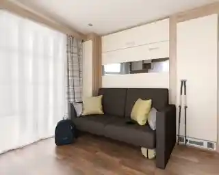 A modern living space featuring a brown sofa with yellow cushions, against a backdrop of light-colored walls and wooden accents. Next to the sofa is a black backpack and a pair of binoculars. Sheer curtains let in natural light, enhancing the cozy ambiance.