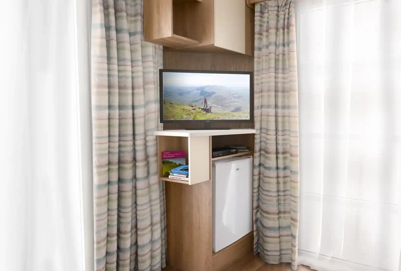 A modern interior featuring an entertainment unit with a TV displaying a scenic landscape. The unit has a shelf with books and a small refrigerator below. Light-colored curtains frame the window, and the flooring is wooden.