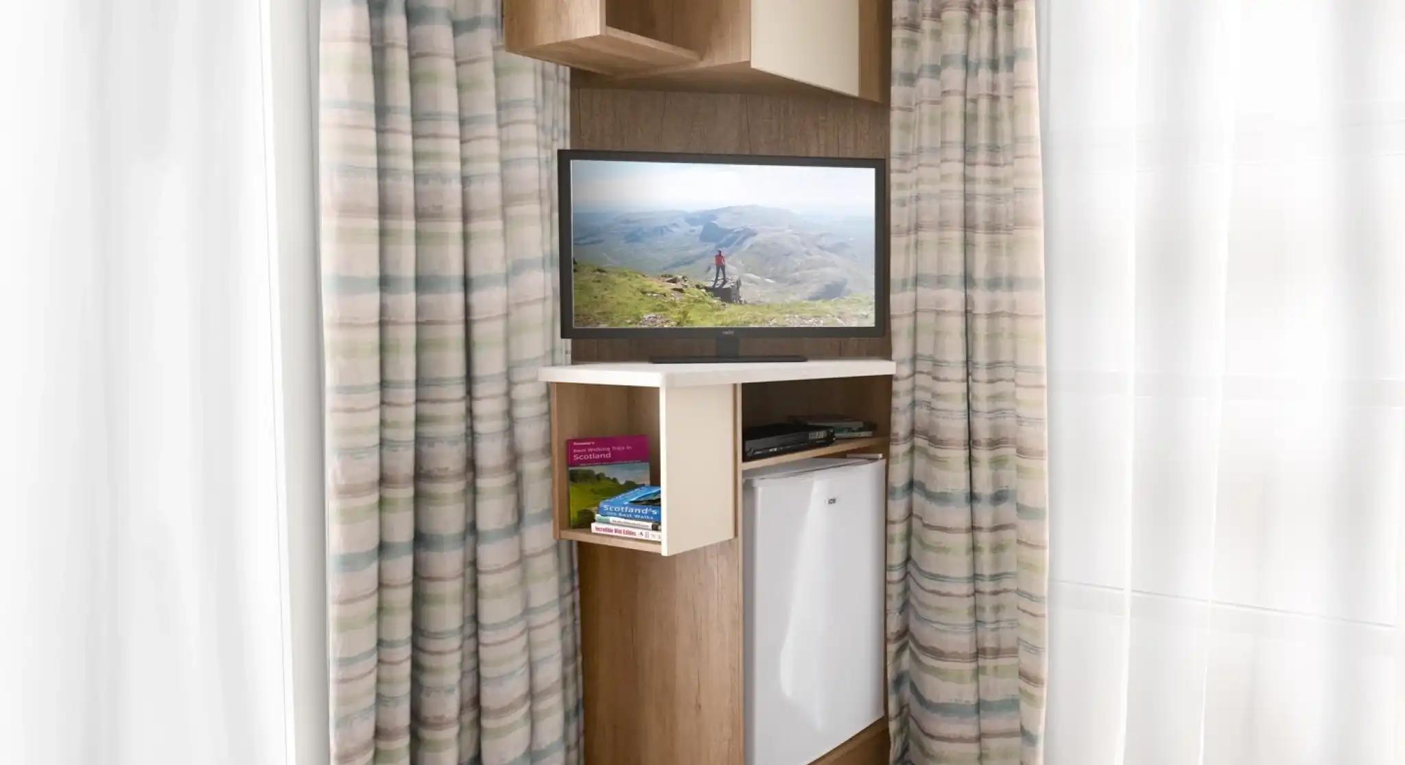 A modern interior featuring an entertainment unit with a TV displaying a scenic landscape. The unit has a shelf with books and a small refrigerator below. Light-colored curtains frame the window, and the flooring is wooden.