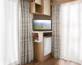 A modern interior featuring an entertainment unit with a TV displaying a scenic landscape. The unit has a shelf with books and a small refrigerator below. Light-colored curtains frame the window, and the flooring is wooden.