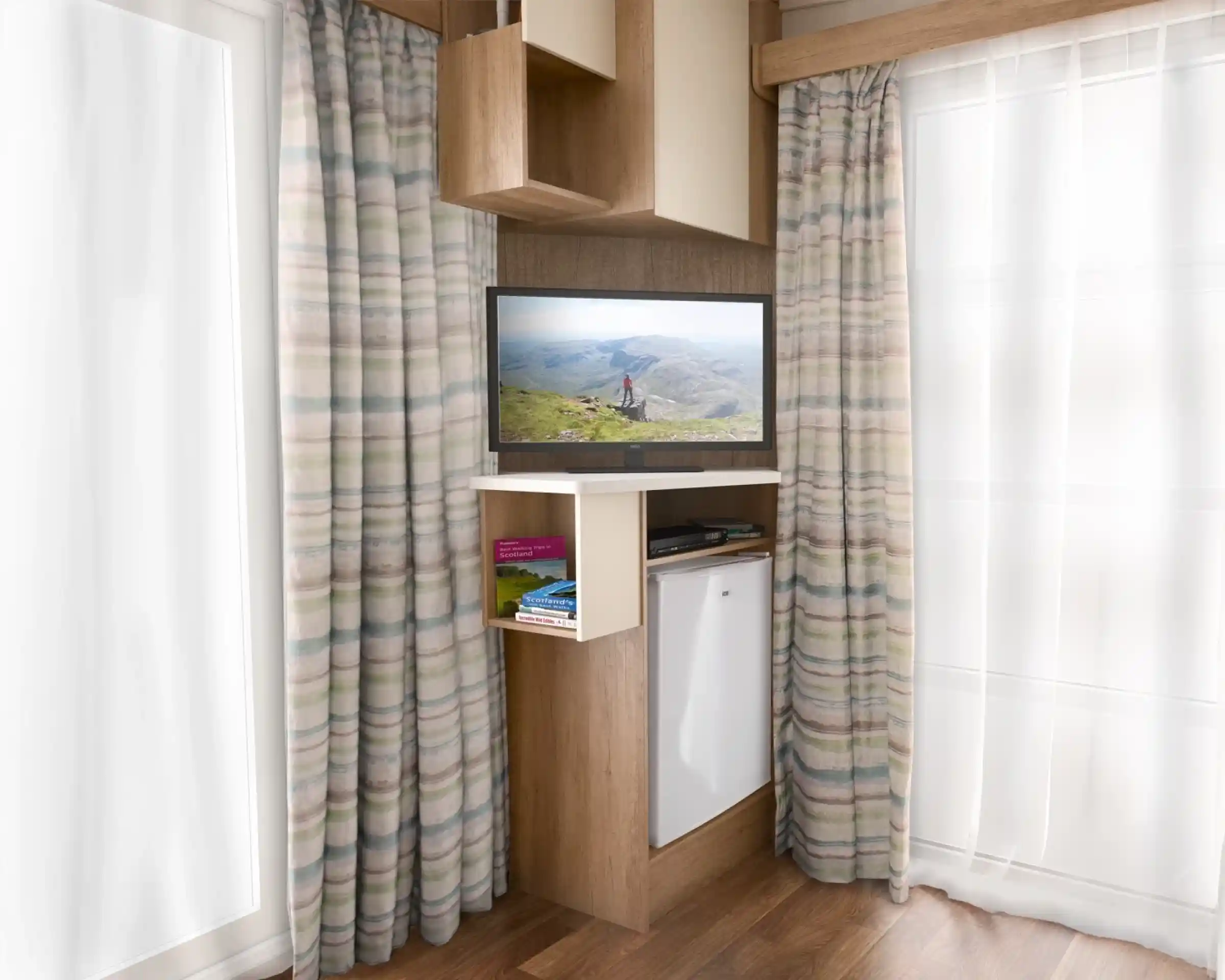 A modern interior featuring an entertainment unit with a TV displaying a scenic landscape. The unit has a shelf with books and a small refrigerator below. Light-colored curtains frame the window, and the flooring is wooden.
