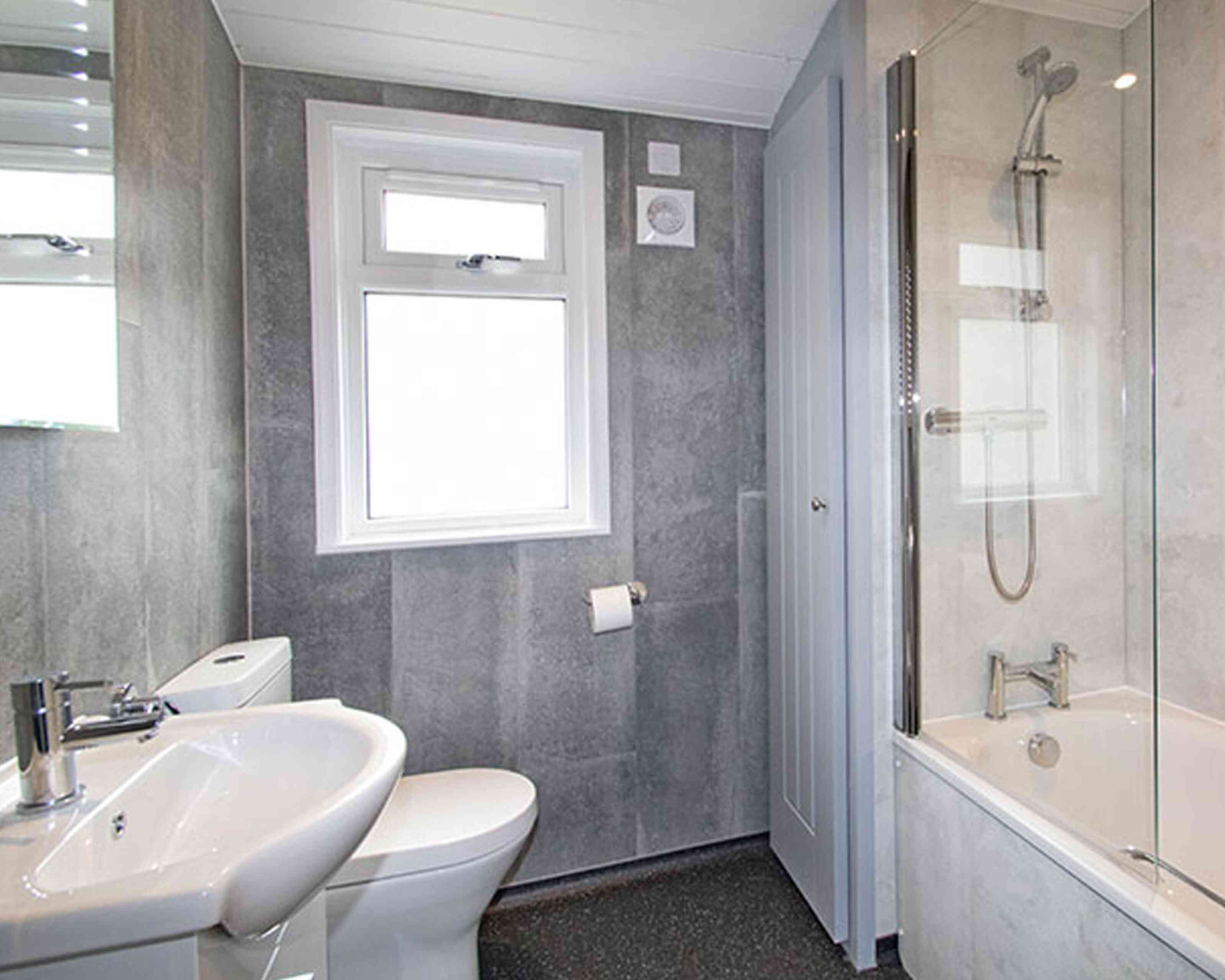 A modern bathroom featuring a gray tiled wall, a white toilet, a sink with a faucet, and a bathtub with a shower. Natural light enters through a window above the sink.