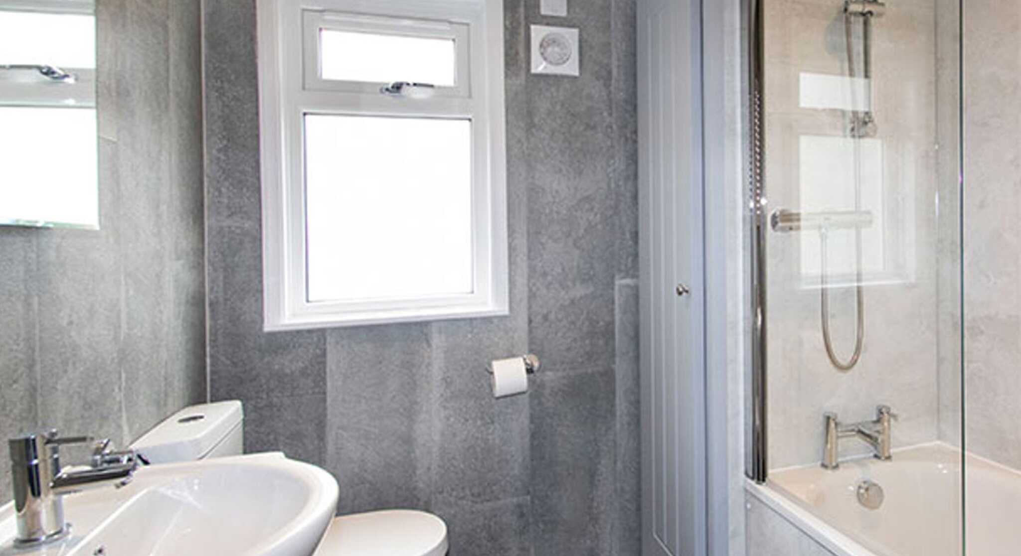 A modern bathroom featuring a gray tiled wall, a white toilet, a sink with a faucet, and a bathtub with a shower. Natural light enters through a window above the sink.