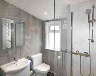 Modern bathroom featuring gray wall tiles, a glass shower enclosure, a sleek sink with a mirror, and a toilet. Natural light enters through a window.