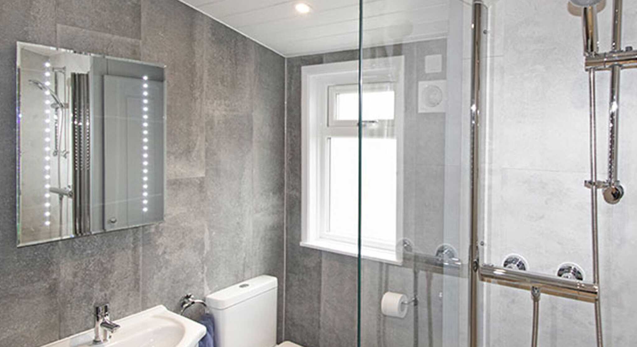 Modern bathroom featuring gray wall tiles, a glass shower enclosure, a sleek sink with a mirror, and a toilet. Natural light enters through a window.
