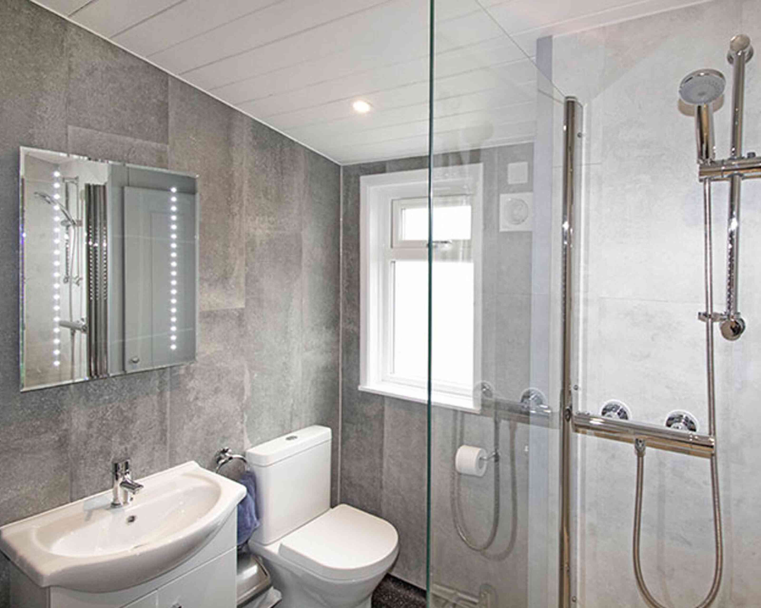 Modern bathroom featuring gray wall tiles, a glass shower enclosure, a sleek sink with a mirror, and a toilet. Natural light enters through a window.