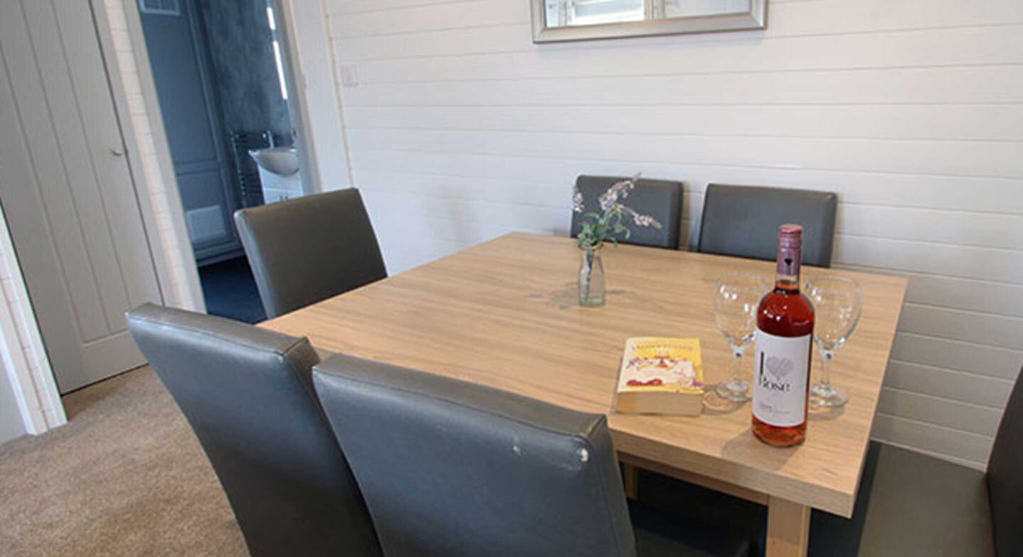 A wooden dining table with four gray chairs is set up in a light, airy room. A bottle of rosé wine, two glasses, and a small vase with flowers are placed on the table. A book lies nearby, and a door can be seen in the background.