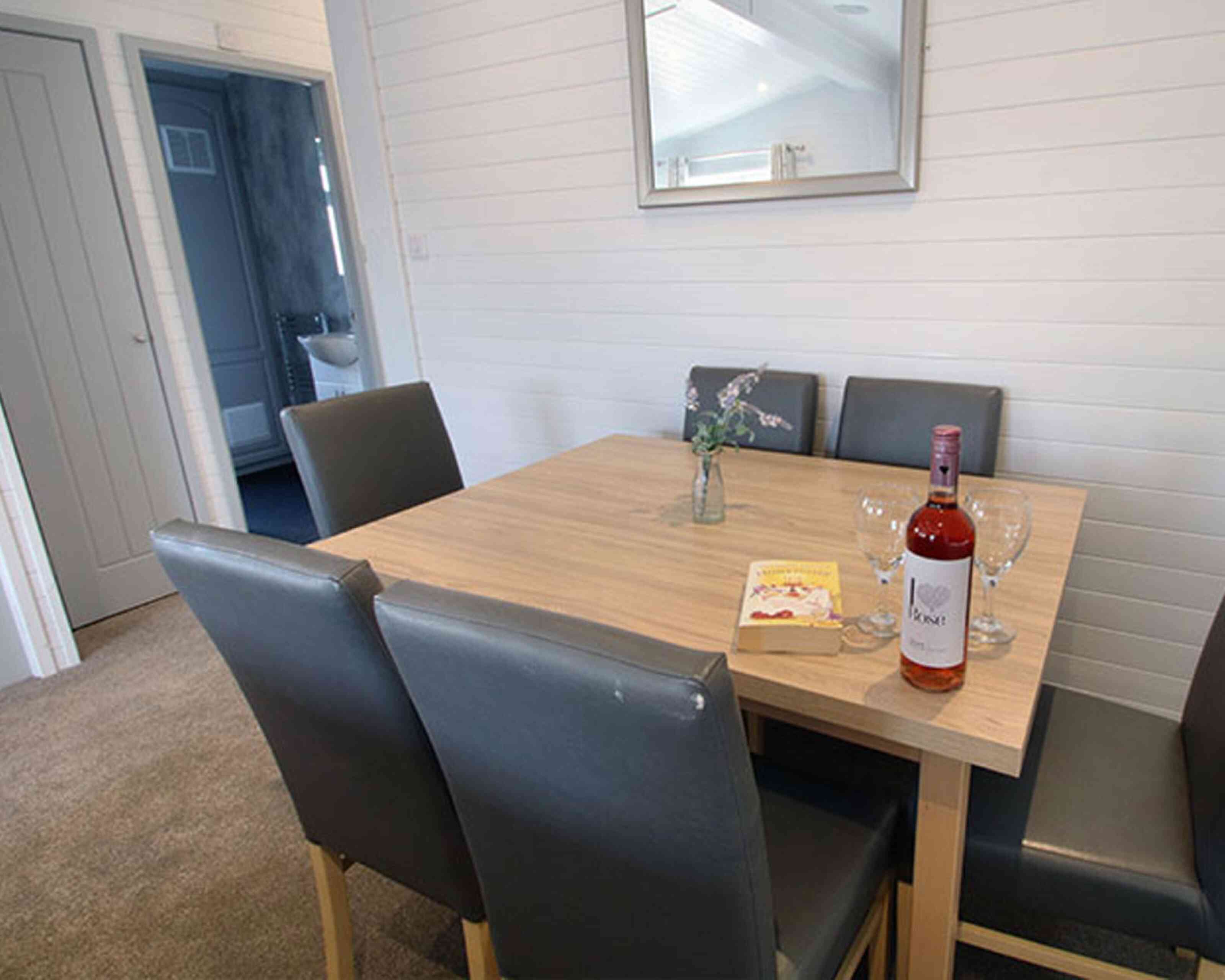 A wooden dining table with four gray chairs is set up in a light, airy room. A bottle of rosé wine, two glasses, and a small vase with flowers are placed on the table. A book lies nearby, and a door can be seen in the background.