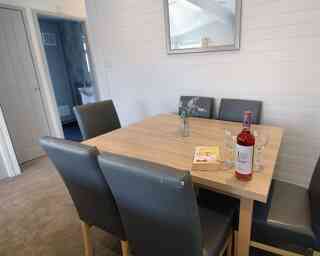 A wooden dining table with four gray chairs is set up in a light, airy room. A bottle of rosé wine, two glasses, and a small vase with flowers are placed on the table. A book lies nearby, and a door can be seen in the background.