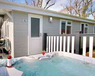 A hot tub with bubbling water is positioned in front of a modern cabin. A bottle of rose wine and a glass sit nearby. The cabin features a white door and a railing with trees in the background.