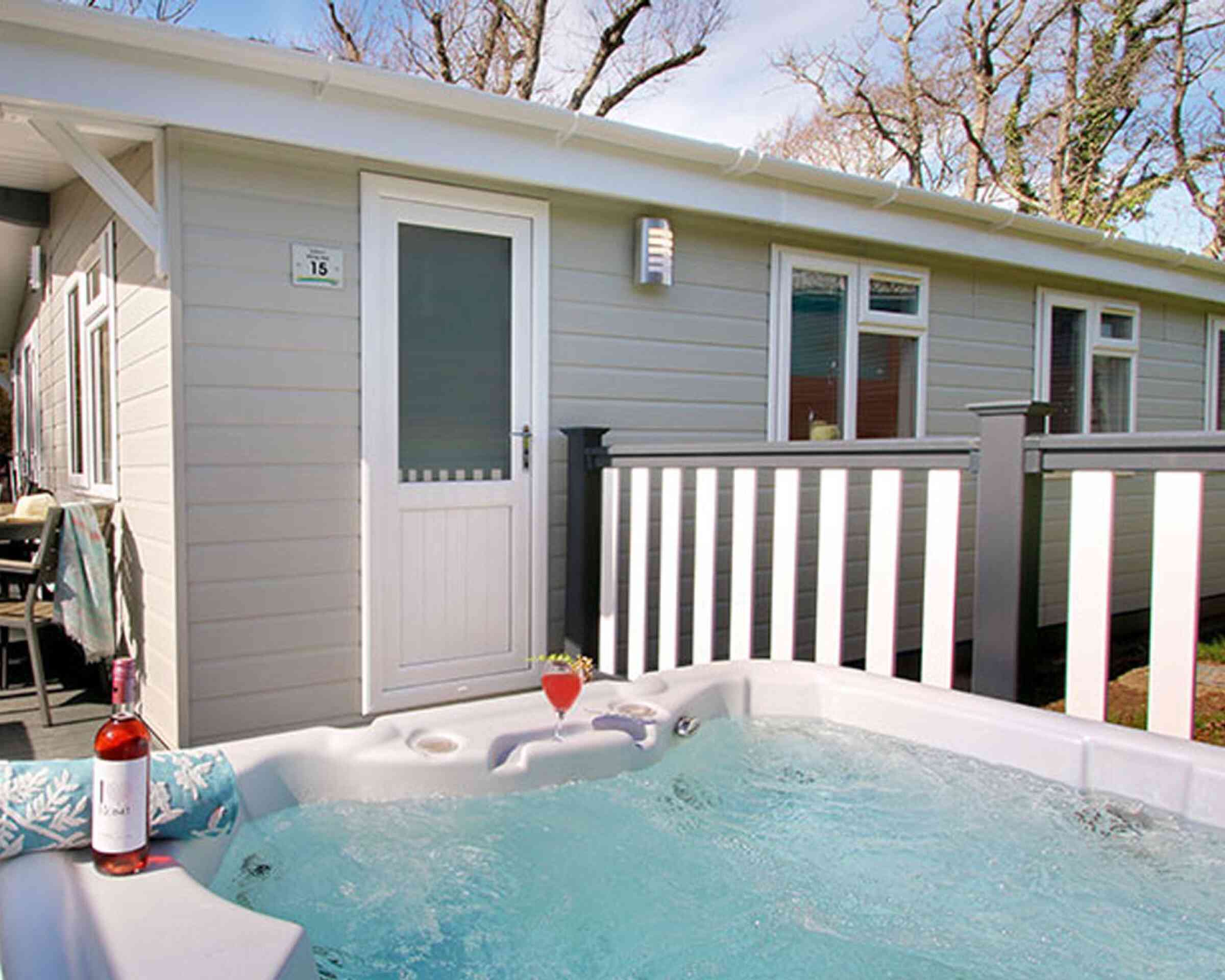 A hot tub with bubbling water is positioned in front of a modern cabin. A bottle of rose wine and a glass sit nearby. The cabin features a white door and a railing with trees in the background.