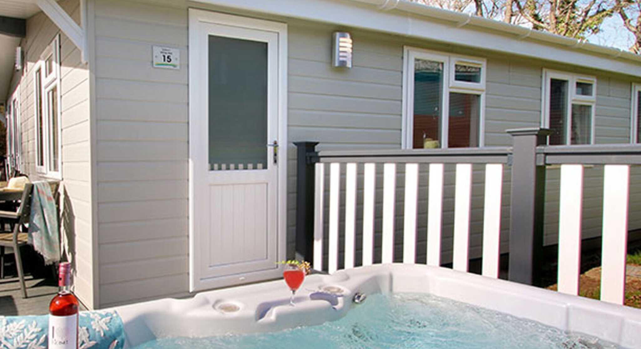 A hot tub with bubbling water is positioned in front of a modern cabin. A bottle of rose wine and a glass sit nearby. The cabin features a white door and a railing with trees in the background.