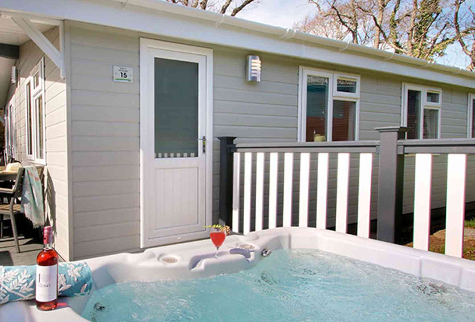 A hot tub with bubbling water is positioned in front of a modern cabin. A bottle of rose wine and a glass sit nearby. The cabin features a white door and a railing with trees in the background.