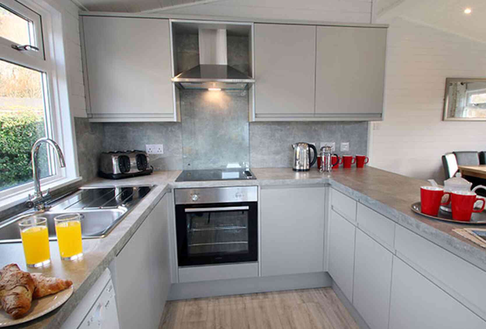 Modern kitchen featuring sleek gray cabinetry, stainless steel appliances, and a breakfast counter with orange juice and pastries. Natural light streams in through a window, illuminating the space.