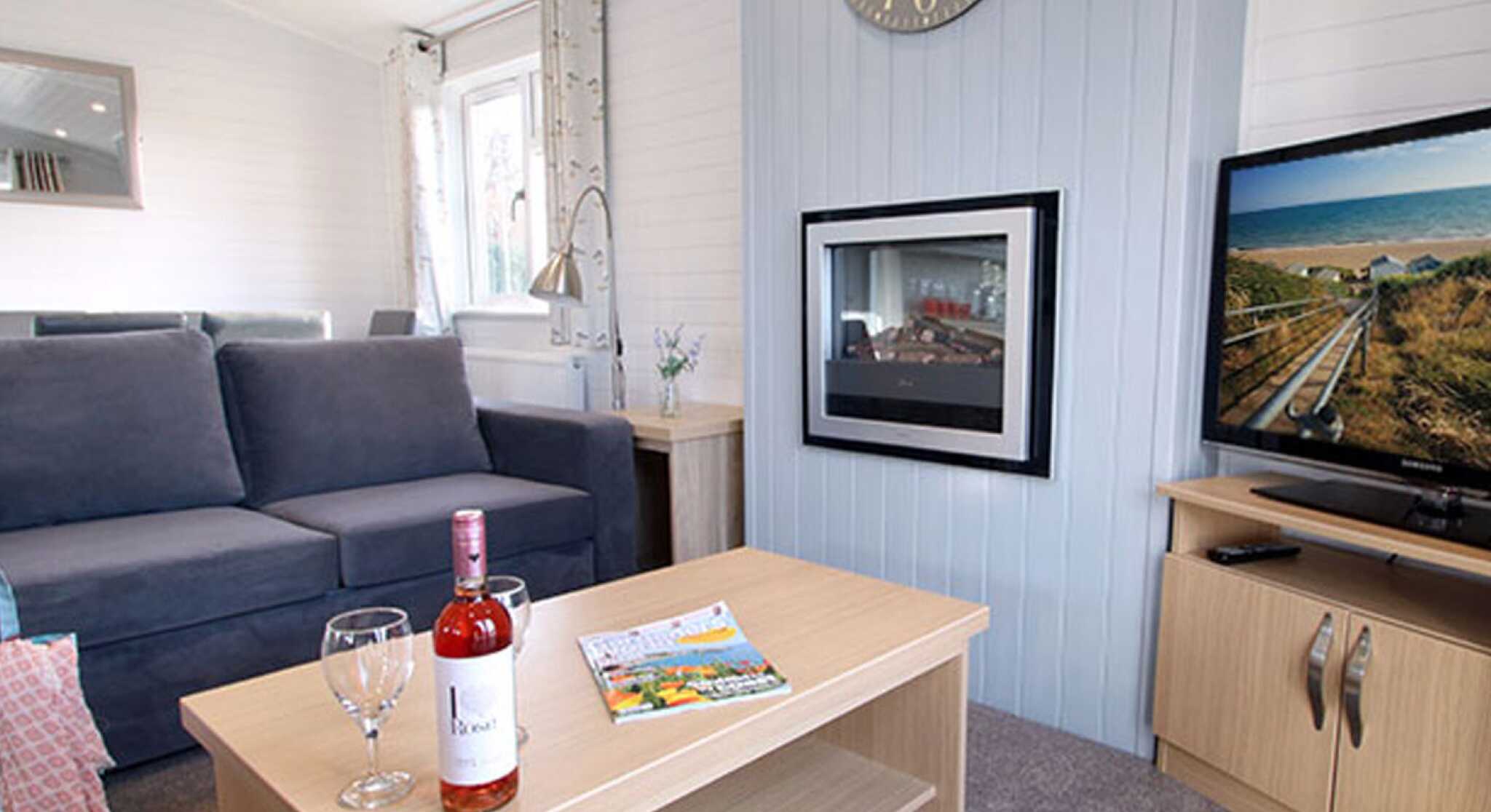A cozy living room featuring a grey sofa, a coffee table with a bottle of rosé wine and two wine glasses, and a television displaying a scenic view. A clock hangs on the wall, and there are large windows allowing natural light.