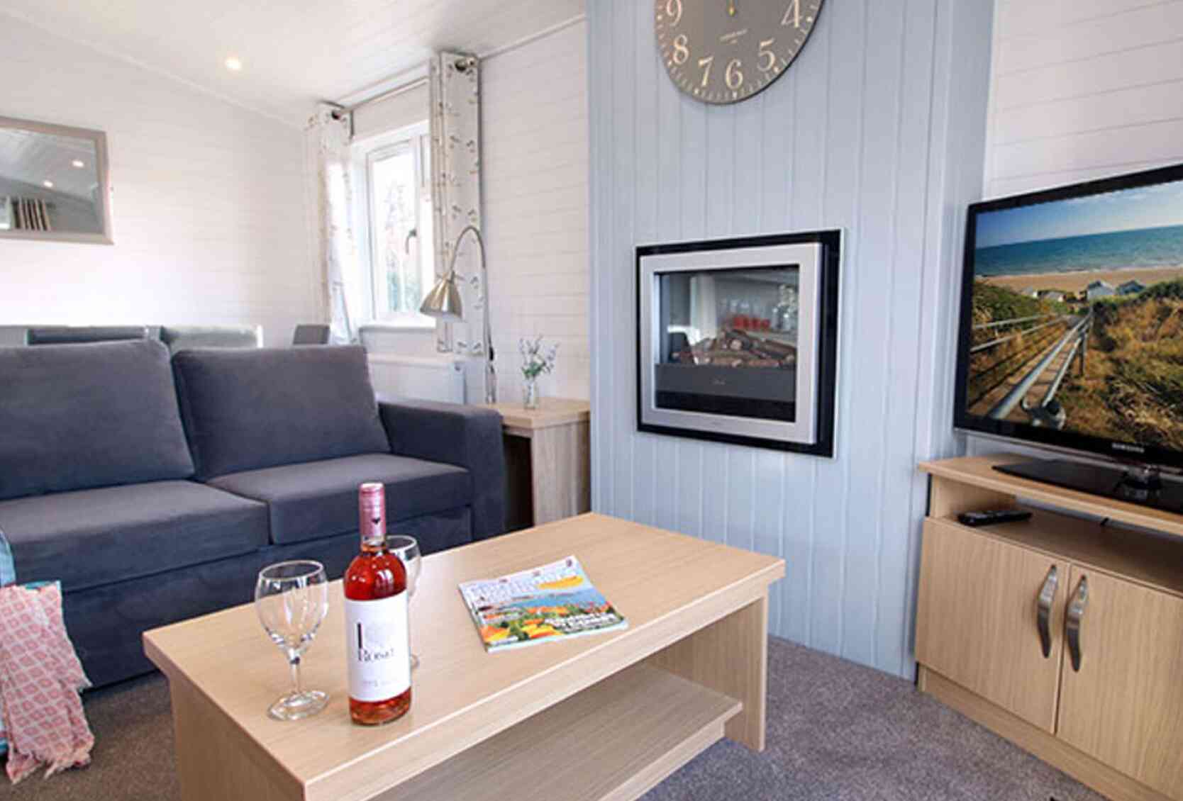 A cozy living room featuring a grey sofa, a coffee table with a bottle of rosé wine and two wine glasses, and a television displaying a scenic view. A clock hangs on the wall, and there are large windows allowing natural light.