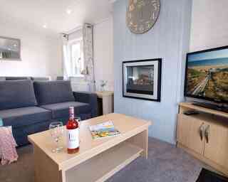 A cozy living room featuring a grey sofa, a coffee table with a bottle of rosé wine and two wine glasses, and a television displaying a scenic view. A clock hangs on the wall, and there are large windows allowing natural light.